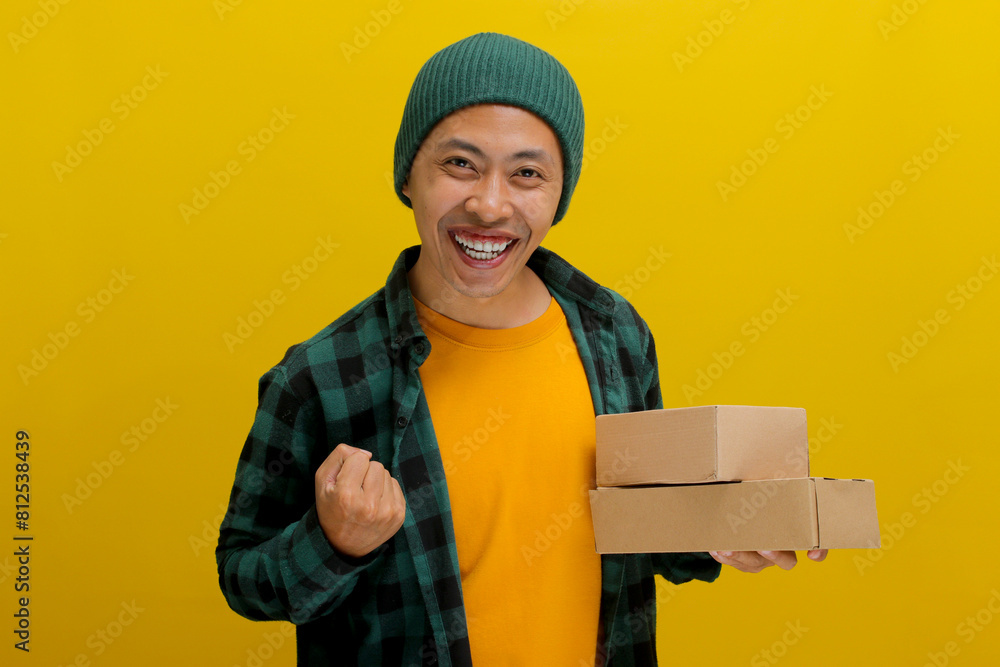 Happy Asian customer joyfully raises his arm in a YES gesture while holding package parcel cardboard box, smiles at the camera, expressing satisfaction upon receiving the awaited online store delivery Happy Asian customer joyfully raises his arm in a YES gesture while holding package parcel cardboard box, smiles at the camera, expressing satisfaction upon receiving the awaited online store delivery