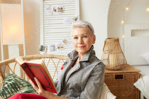 Indoor portrait of beautiful caucasian senior woman in trendy casual clothes looking at camera while holding red hardcover book of novel, enjoying leisure time sitting in wicker chair