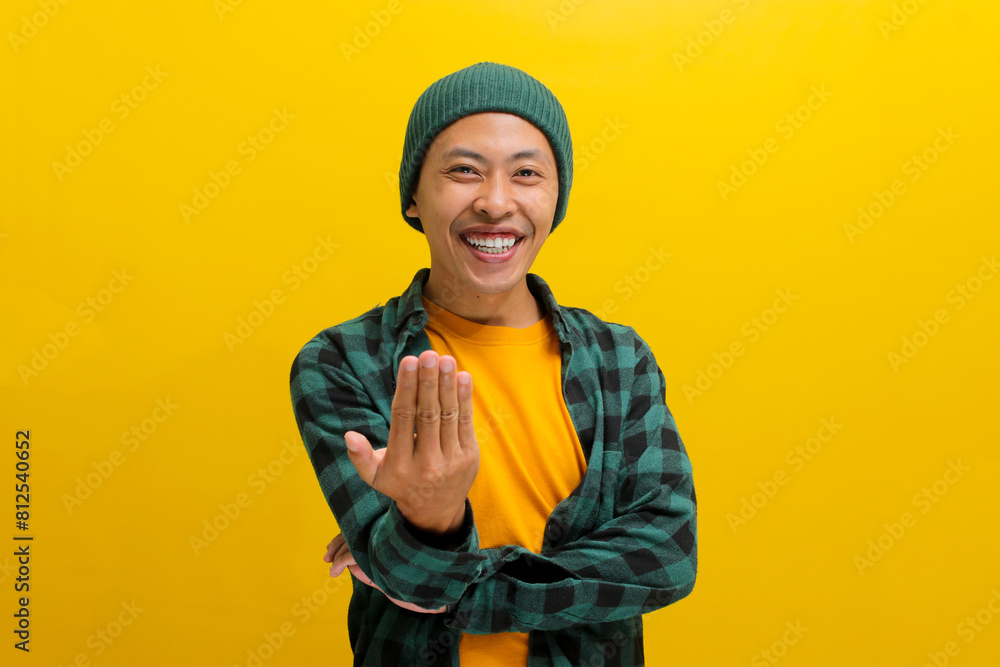 Happy, smiling young Asian man, dressed in a beanie hat and casual shirt, invites with a beckoning COME HERE gesture while standing against a yellow background. Happy, smiling young Asian man, dressed in a beanie hat and casual shirt, invites with a beckoning COME HERE gesture while standing against a yellow background.