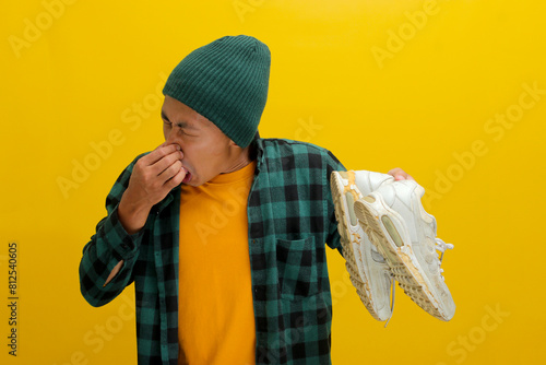 Asian man in a beanie and plaid shirt grimaces while pinching his nose shut, likely reacting to a strong odor. Isolated on a yellow background. Unpleasant smells, disgust, and humor concept photo