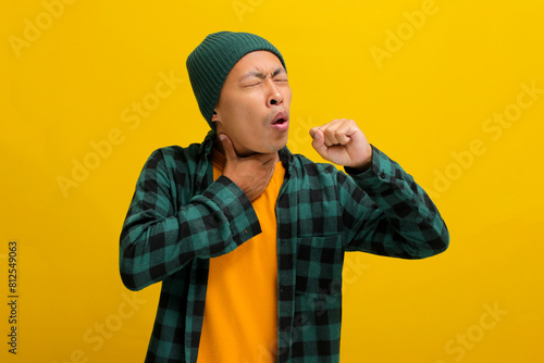 Young Asian man, wearing a beanie hat and casual shirt, appears to be experiencing throat pain and dry coughing, possibly due to a virus or infection, isolated on a yellow background.