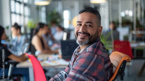 Smiling man with beard in plaid shirt sitting in modern office with blurred colleagues in background.