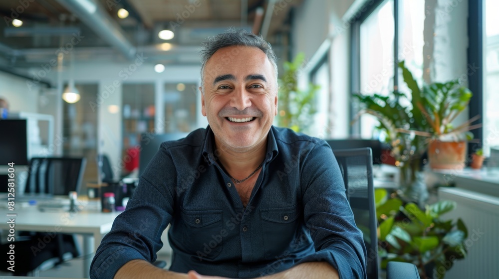 Smiling man wearing blue shirt sitting in office chair with plants in background. Smiling man wearing blue shirt sitting in office chair with plants in background.