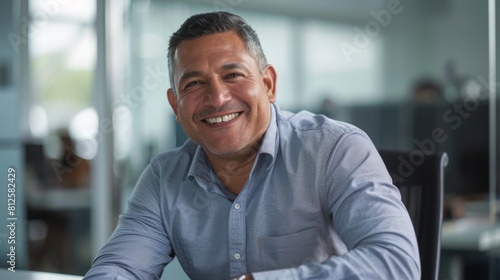 Smiling man in office wearing blue shirt sitting at desk with blurred background. © iuricazac
