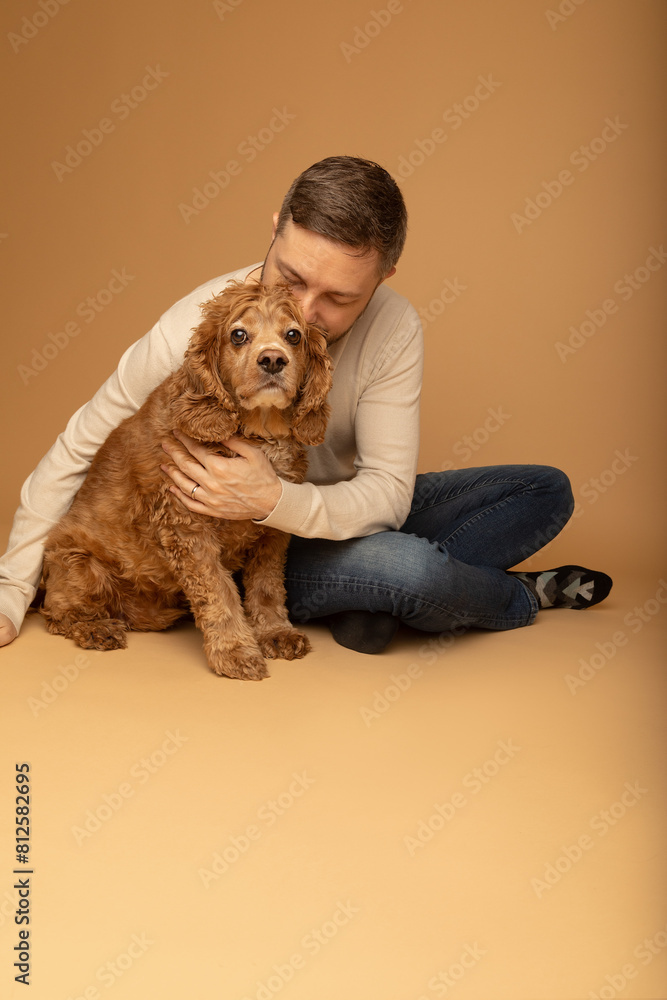 Obraz premium Studio portrait of a handsome caucasian man in his 40s sitting on the floor and embracing his cocker spaniel dog. The background is beige. The man is wearing jeans and beige shirt. 