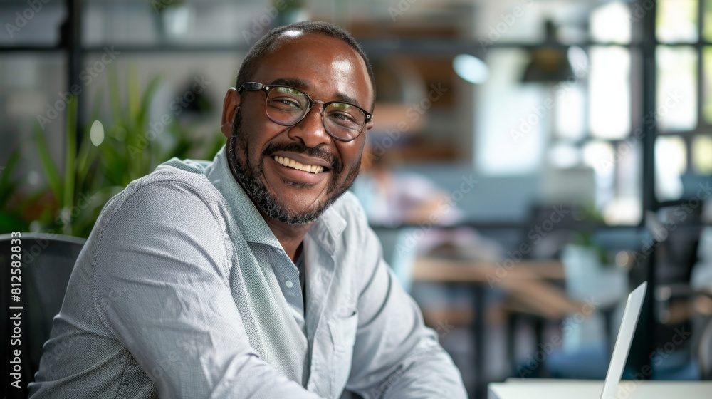 Smiling man in glasses wearing a light blue shirt sitting at a table with a laptop in a modern well-lit indoor setting with blurred background. Smiling man in glasses wearing a light blue shirt sitting at a table with a laptop in a modern well-lit indoor setting with blurred background.
