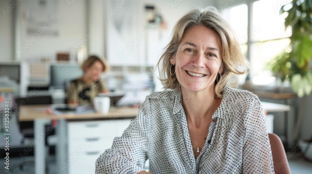 Smiling woman in office setting wearing patterned blouse seated at desk with blurred background of co-worker and office environment. Smiling woman in office setting wearing patterned blouse seated at desk with blurred background of co-worker and office environment.
