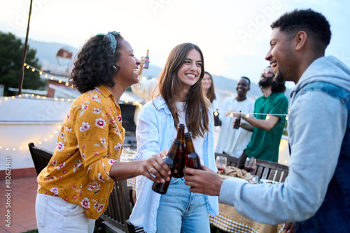 Young group of multi ethnic young people dancing together and toasting beer at rooftop party outside. Mixed race happy friends having fun cheering drink while celebrating birthday at home terrace. © CarlosBarquero