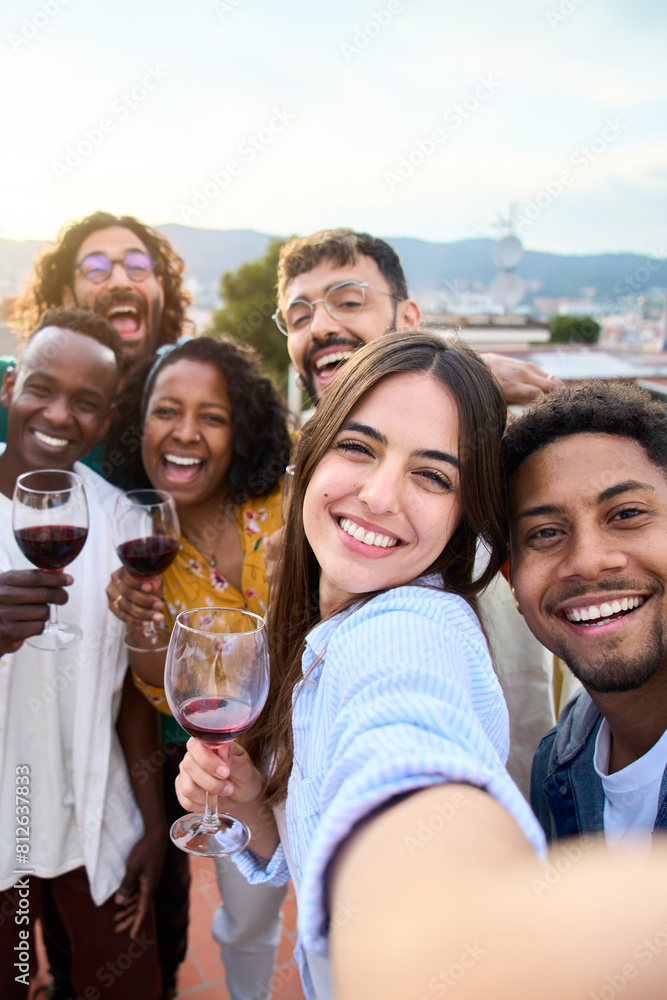Obraz premium Group of diverse friends taking selfie and having fun at rooftop home party holding glass of red wine. Beautiful young people. Smiling community drinking on outdoor terrace. Positive friendship