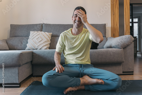 Man on yoga block in living room with legs crossed and a hand in his forehead reacting playfully with laughter and surprised gesture before guided meditation.