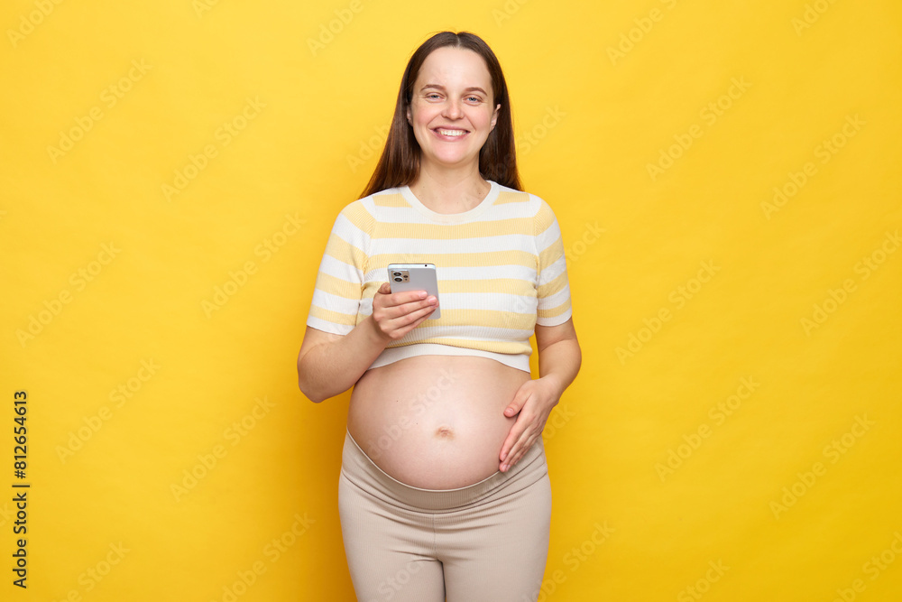 Delighted pretty happy brown haired pregnant woman with bare belly posing isolated over yellow background using mobile phone looking at camera with toothy smile Delighted pretty happy brown haired pregnant woman with bare belly posing isolated over yellow background using mobile phone looking at camera with toothy smile