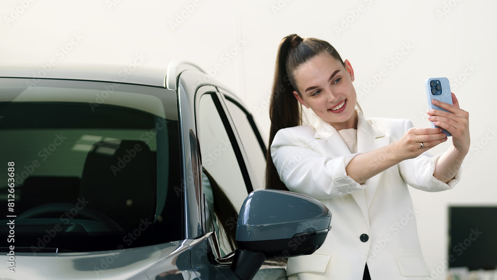 A beautiful female client made a purchase of an electric car in a modern car dealership. A beautiful woman reports her purchase on her phone and takes a selfie. The concept of an expensive purchase, m A beautiful female client made a purchase of an electric car in a modern car dealership. A beautiful woman reports her purchase on her phone and takes a selfie. The concept of an expensive purchase, m