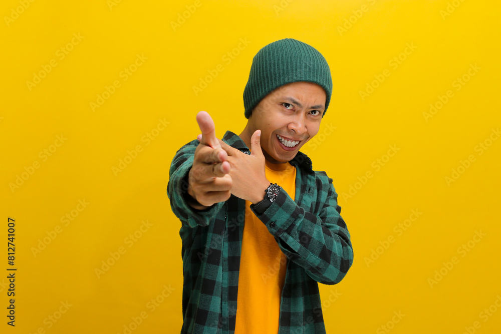 Confident young Asian man, points at the camera with a finger gun gesture, displaying confidence, making choices, and aiming with hand pistol gesture right on target while standing on yellow backdrop Confident young Asian man, points at the camera with a finger gun gesture, displaying confidence, making choices, and aiming with hand pistol gesture right on target while standing on yellow backdrop