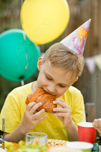Cute funny nine year old boy celebrating his birthday with family or friends and eating croissant in a backyard. Birthday party for kids.