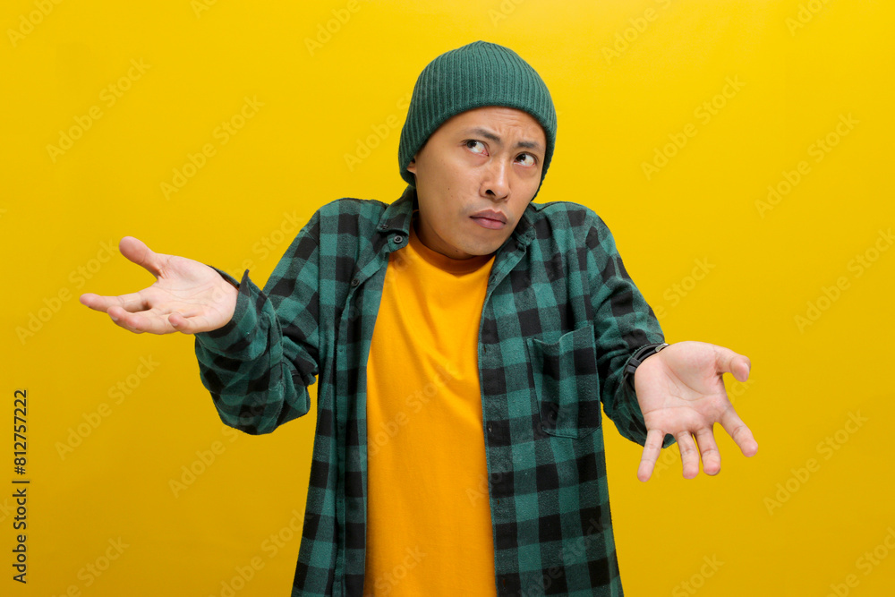 Confused young Asian man, dressed in a beanie hat and casual shirt is shrugging his shoulders in a gesture of cluelessness while standing against yellow background. Confused young Asian man, dressed in a beanie hat and casual shirt is shrugging his shoulders in a gesture of cluelessness while standing against yellow background.