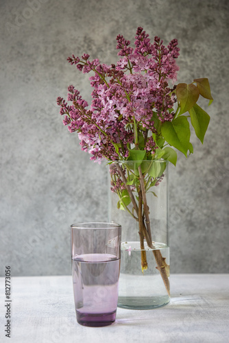 Fresh picked lilac branches in transparent glass vase with a purple glass. Grey background. Copy space.