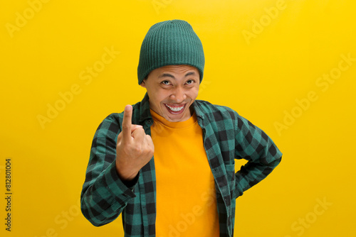 Happy, smiling young Asian man, dressed in a beanie hat and casual shirt, invites with a beckoning COME HERE gesture while standing against a yellow background. © Jamaludinyusup