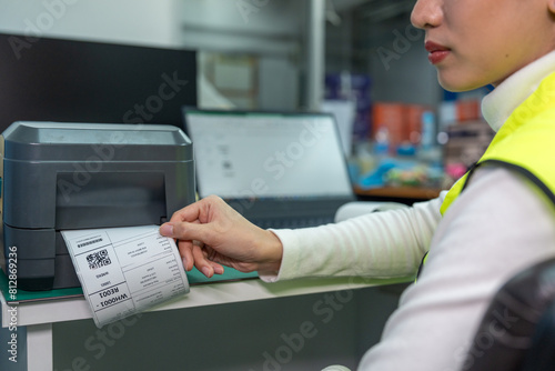 Beautiful Asian woman uses a laptop or notebook to print bar code stickers on a bar code printer. Selective focus at sticker barcode.