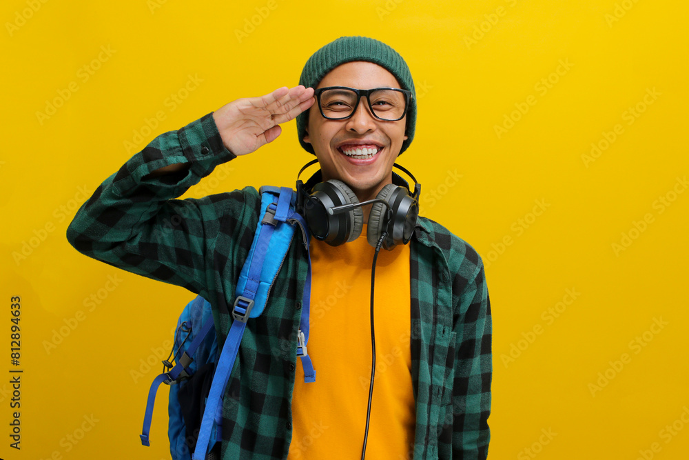 Fototapeta premium A young Asian man, dressed in a beanie hat and casual clothes, salutes with a cheerful expression and a smile directed at the camera while standing against a yellow background