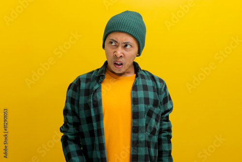 Displeased young Asian man, dressed in a beanie hat and casual shirt, is clearly expressing his disgust at something awful while standing against yellow background. photo