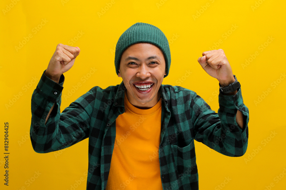 Excited young Asian man, dressed in a beanie hat and casual shirt, is expressing joy and celebrating victory with a fist pump or a YES gesture while standing against a yellow background Excited young Asian man, dressed in a beanie hat and casual shirt, is expressing joy and celebrating victory with a fist pump or a YES gesture while standing against a yellow background