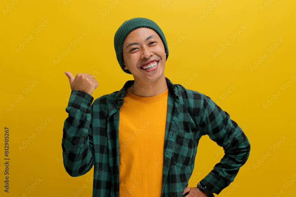 An excited young Asian man, wearing a beanie hat and casual shirt, is smiling while pointing with his thumb to an empty space beside him against a yellow background An excited young Asian man, wearing a beanie hat and casual shirt, is smiling while pointing with his thumb to an empty space beside him against a yellow background