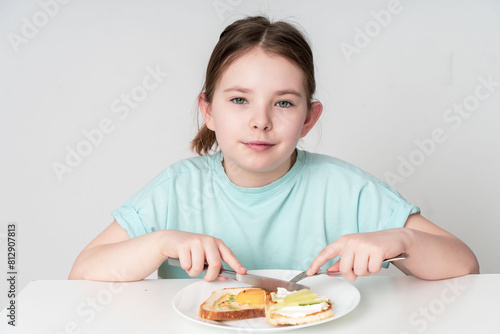 A cute little girl has breakfast with bruschetta with egg, cucumber and cottage cheese. Teenage girl eating healthy breakfast on white background. The concept of healthy eating. photo