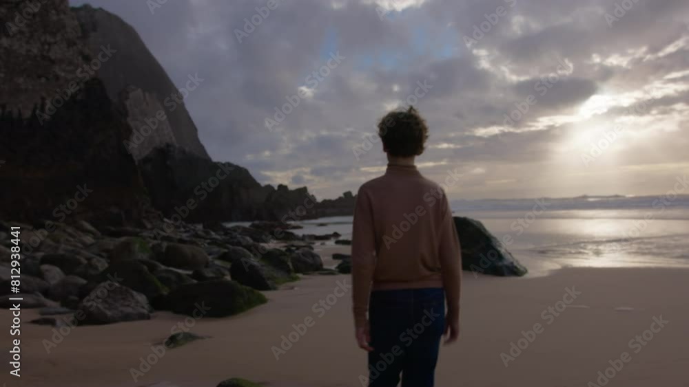 A teenage boy in a brown sweater and jeans strolls along a sandy beach with large rocks A teenage boy in a brown sweater and jeans strolls along a sandy beach with large rocks