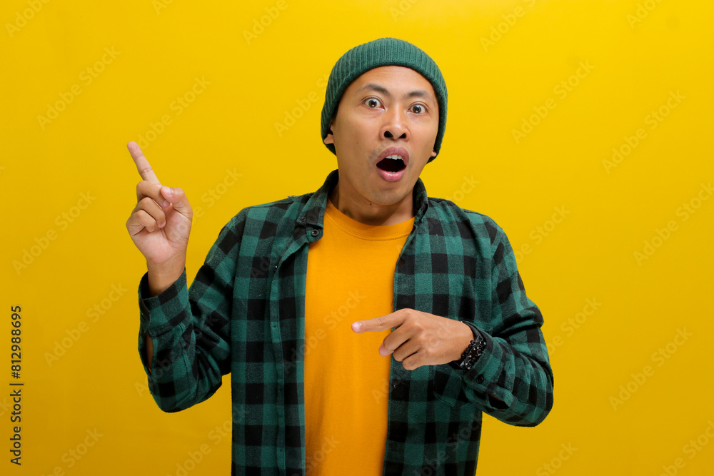 Astonished Asian man, dressed in a beanie hat and casual shirt, points at an empty space, possibly promoting a product or sharing information about an incredible offer, isolated on yellow background. Astonished Asian man, dressed in a beanie hat and casual shirt, points at an empty space, possibly promoting a product or sharing information about an incredible offer, isolated on yellow background.