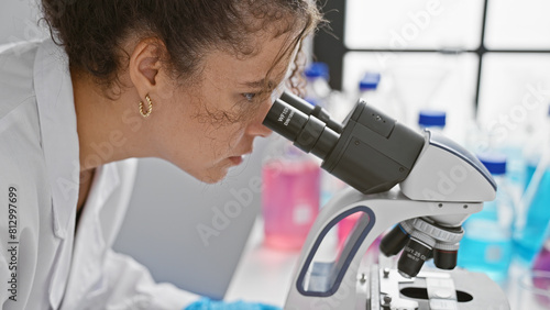 A young hispanic woman with curly hair examines samples through a microscope in a laboratory setting.