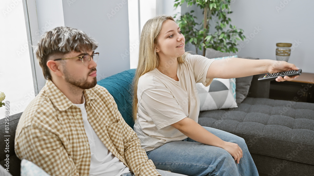 A woman changing tv channels next to a man on a couch in a modern living room.