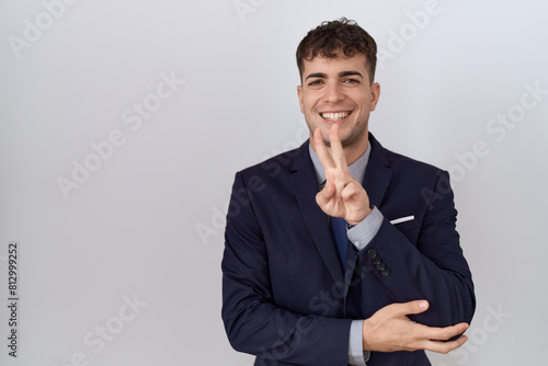 Young hispanic business man wearing suit and tie smiling with happy face winking at the camera doing victory sign. number two.