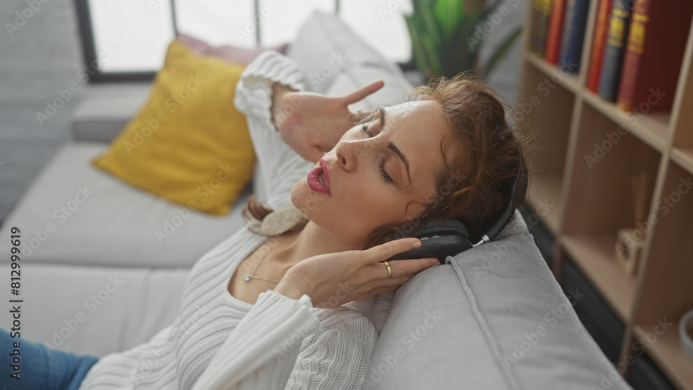 A relaxed young woman enjoying music with headphones while lying on a couch in a cozy living room. A relaxed young woman enjoying music with headphones while lying on a couch in a cozy living room.