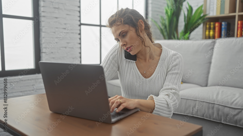 A multitasking young woman works on her laptop with a phone held between shoulder and ear in a bright home interior.