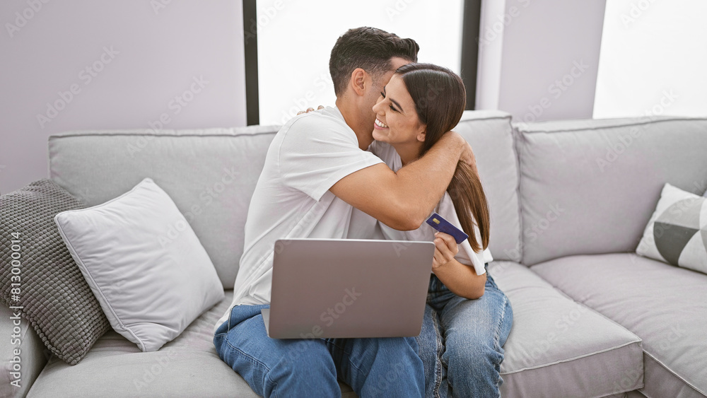 A loving couple cuddles on a sofa in a modern living room, with the woman holding a credit card and a laptop in the background.