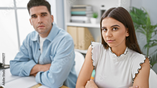 A serious man and woman with arms crossed sit in a modern office  displaying teamwork in a corporate environment.
