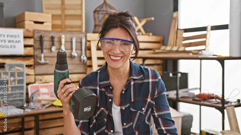 A smiling woman in safety glasses holds a drill in a well-equipped carpentry workshop. A smiling woman in safety glasses holds a drill in a well-equipped carpentry workshop.