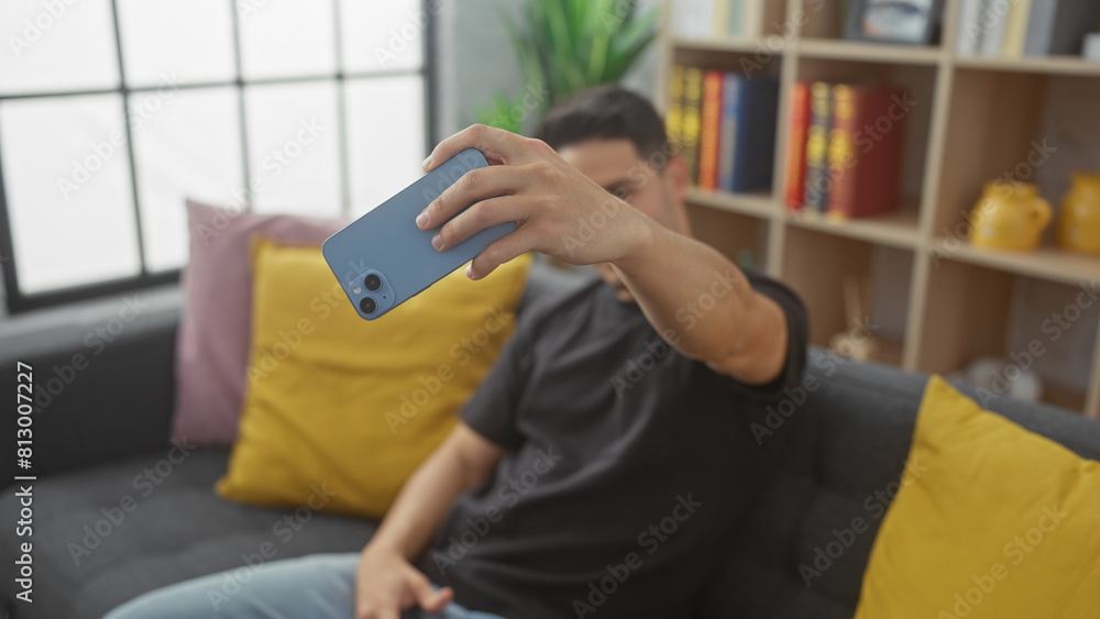 Obraz premium Hispanic man takes selfie with smartphone in a cozy home living room.