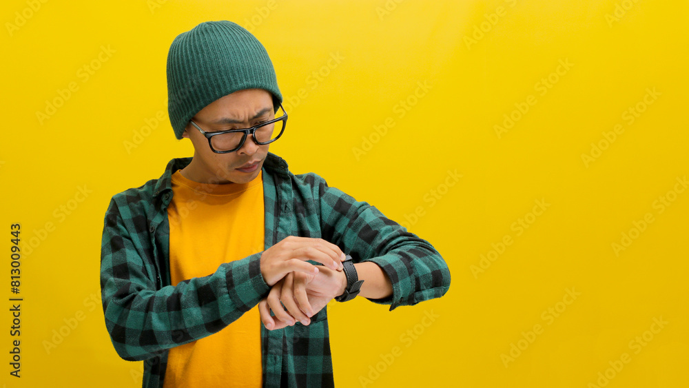Asian man in a beanie and casual shirt checks the time on his wristwatch, isolated on a yellow background Asian man in a beanie and casual shirt checks the time on his wristwatch, isolated on a yellow background