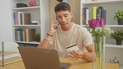 Hispanic man multitasking with phone and credit card at home office setup by laptop and flowers.