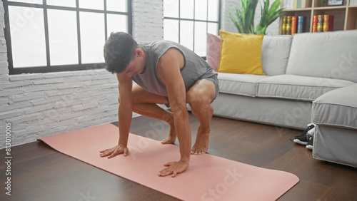 A young man doing a burpee on a pink exercise mat in a bright living room with a gray sofa and large windows.