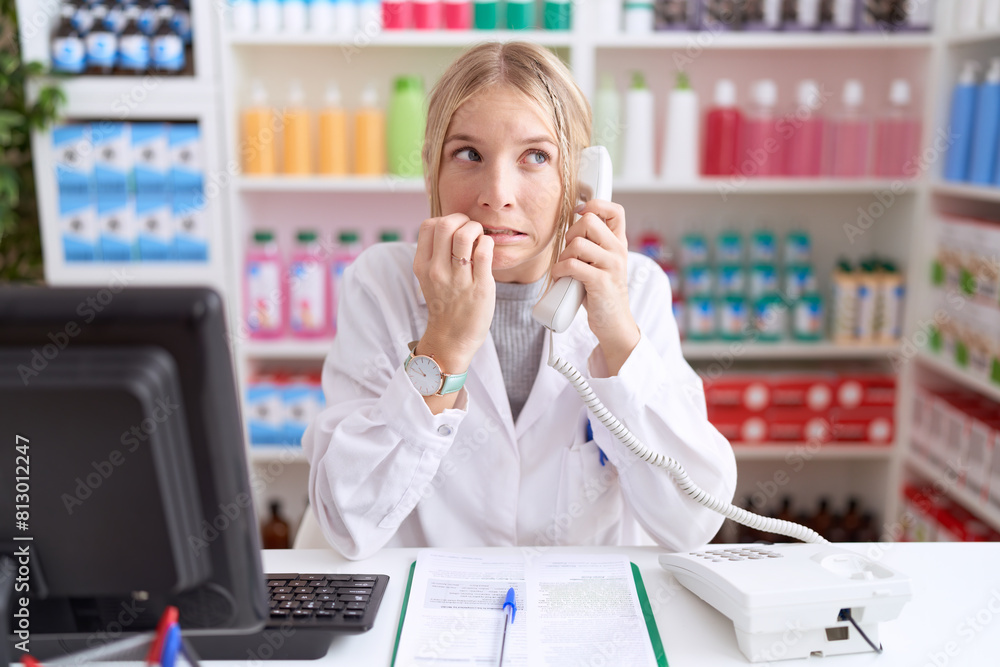 Young caucasian woman working at pharmacy drugstore speaking on the telephone looking stressed and nervous with hands on mouth biting nails. anxiety problem. Young caucasian woman working at pharmacy drugstore speaking on the telephone looking stressed and nervous with hands on mouth biting nails. anxiety problem.