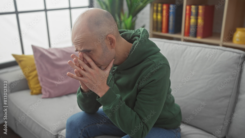A concerned bald man with a beard sitting on a couch indoors looking stressed, covering his face with his hands. A concerned bald man with a beard sitting on a couch indoors looking stressed, covering his face with his hands.