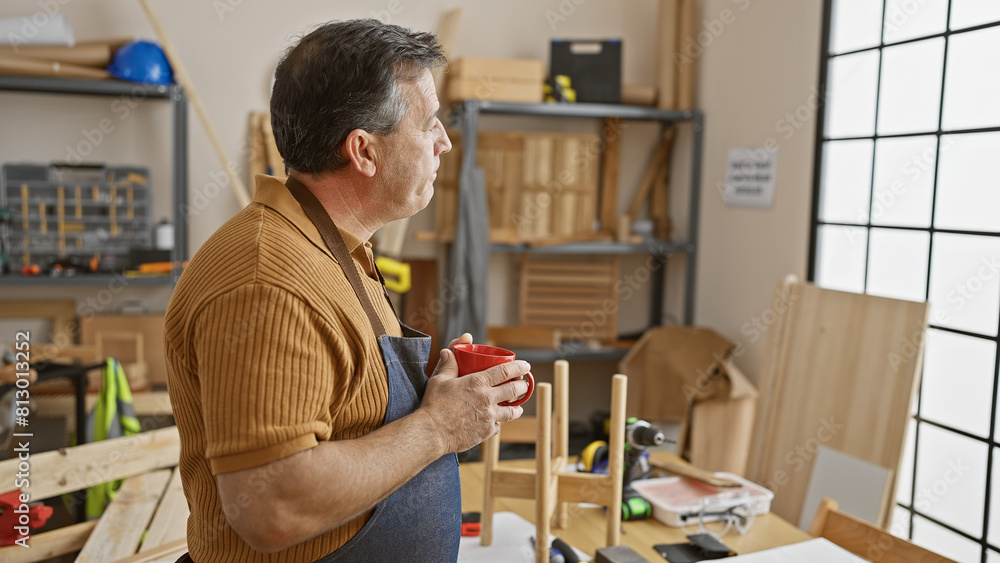 A mature man in an apron stands contemplatively with a mug in a well-equipped woodworking workshop.