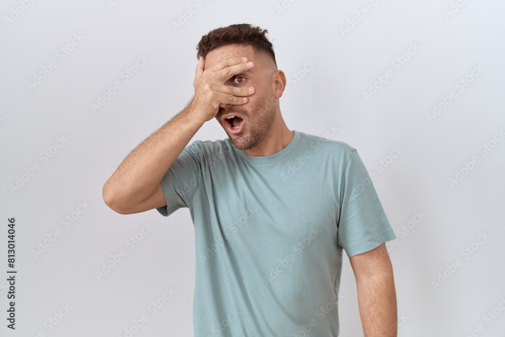 Hispanic man with beard standing over white background peeking in shock covering face and eyes with hand, looking through fingers with embarrassed expression. Hispanic man with beard standing over white background peeking in shock covering face and eyes with hand, looking through fingers with embarrassed expression.