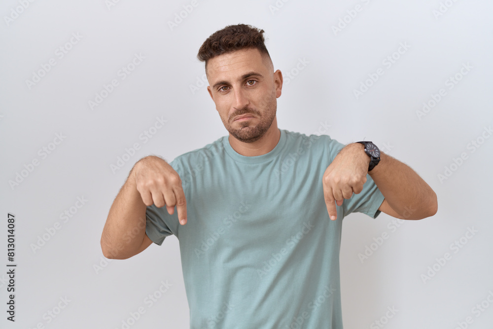 Hispanic man with beard standing over white background pointing down looking sad and upset, indicating direction with fingers, unhappy and depressed. Hispanic man with beard standing over white background pointing down looking sad and upset, indicating direction with fingers, unhappy and depressed.