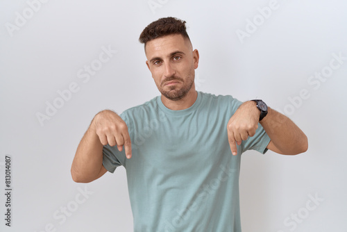 Foto Hispanic man with beard standing over white background pointing down looking sad and upset, indicating direction with fingers, unhappy and depressed