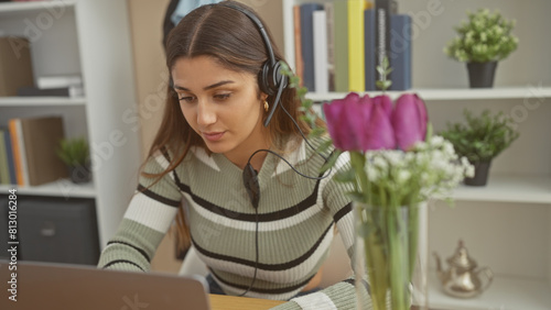 A young latina woman works on a laptop at home, wearing headphones and a striped sweater, focused and serene.