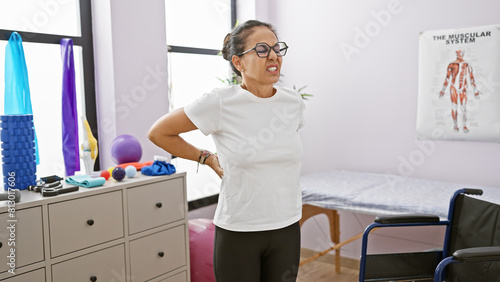 Hispanic senior woman with curly hair experiencing back pain at a physiotherapy clinic interior.