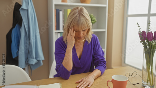 A stressed woman sits at a table in her home office with glasses, paperwork, a cup, and flowers, conveying domestic work-life balance.